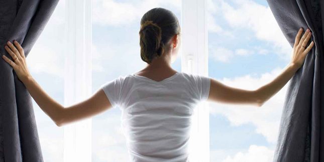 Foto: Initiative Gute Luft - Frau öffnet Vorhänge und schaut in die Natur. Wohnraumlüftung ermöglicht Allergikern, das ganze Jahr frei durchzuatmen. 