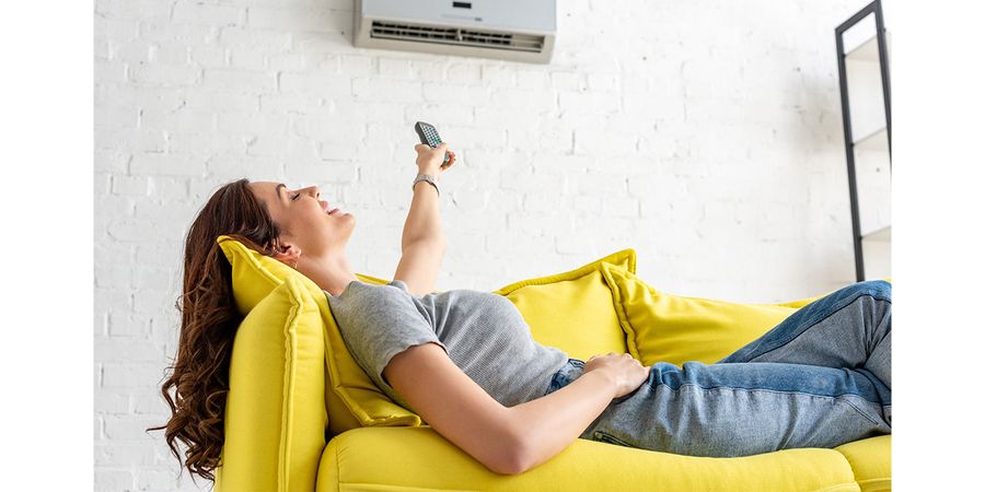 Klimaanlage zuhause: Frau sitzt gemütlich auf der Couch und bedient die Klimaanlage mit einer Fernbedienung