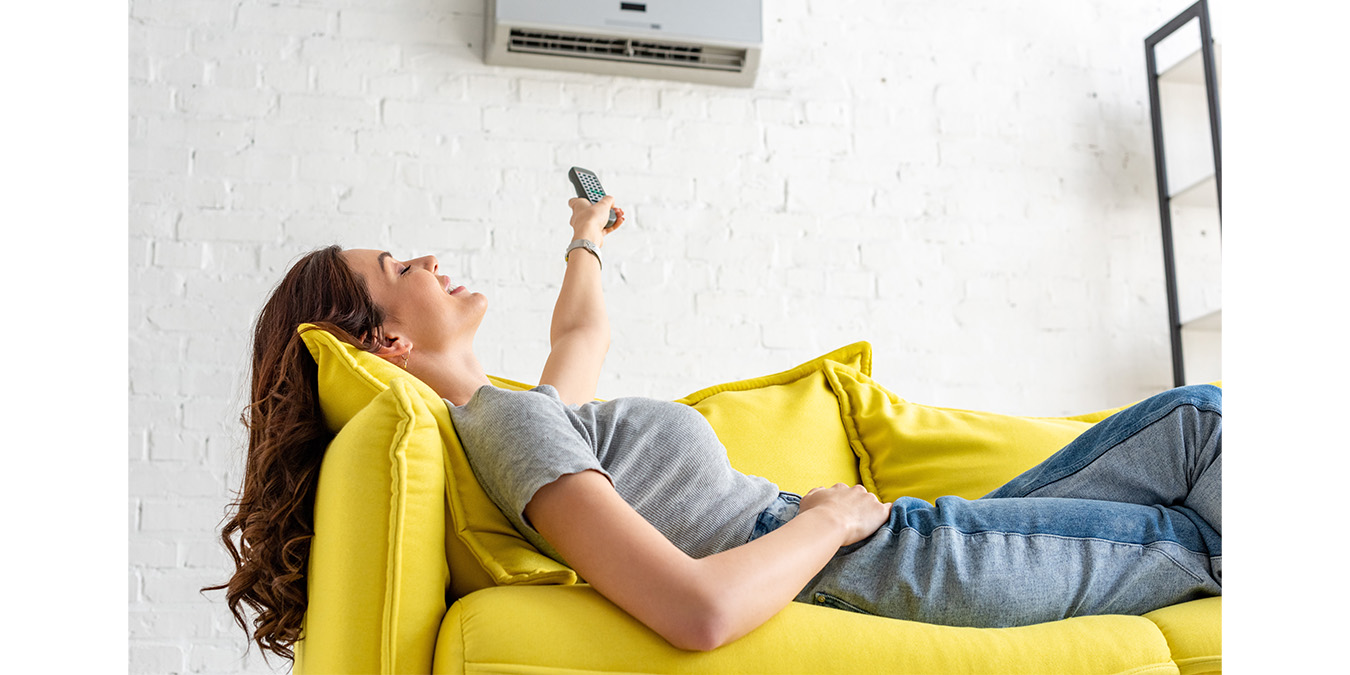 Klimaanlage zuhause: Frau entspannt auf der Couch, hat Fernbedienung in der Hand und schaltet die Klimaanlage ein